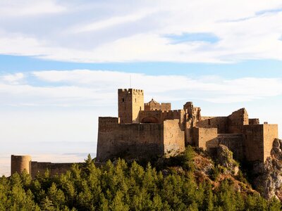 Castle of Loarre on sunny day viewed from the distance with pine trees in foreground, and cloudy blue skies in background, Spain
