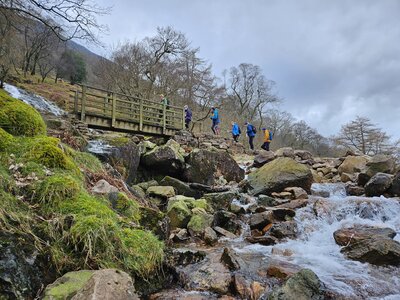 Ramble Worldwide walking holiday navigation & hill walking skills group crossing bridge over stream from waterfall nearby, Buttermere, Lake District, Cumbria