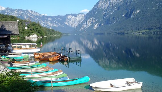 Small boats docked up at shore with mountains in distance reflecting in Lake Bohinj on sunny day, Slovenia